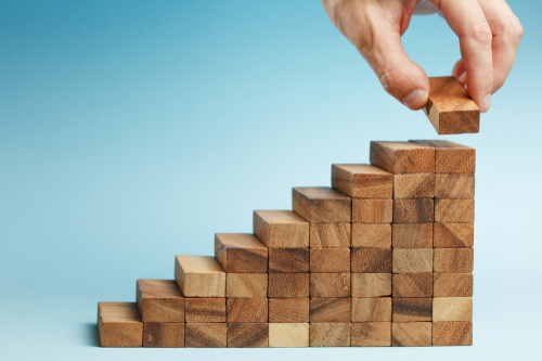 Ein Hand, die die letzte Stufe einer Treppe aus Holz-DominoSteinen legt.