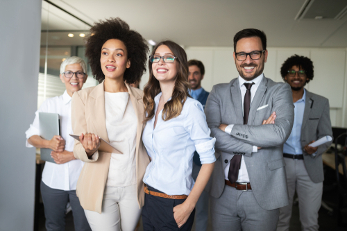 Eine Gruppe von Büro-Mitarbeitern verschiedenster Altersstufen und Abstammung die einen zufrieden und fröhlich anschauen.