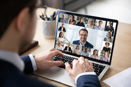 Ein locker gekleideter Anzugträger, der an seinem Laptop am Tisch sitzt. Auf dem Laptop-Bildschirm ist bereits die Gesichter aller seiner Kollegen zu sehen, dies sich gerade im Online-Meeting treffen.